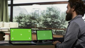 Wealthy person using greenscreen on two devices, sitting at his workstation in his modern elegant vacation home. Rich business owner and investor looking at screens running blank chromakey. photo