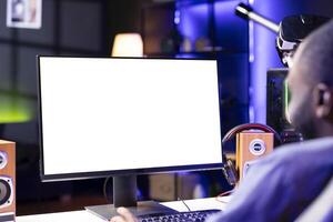 African american man in dimly lit apartment playing videogames on isolated screen gaming PC at computer desk, enjoying day off from work. BIPOC person enjoying game on mockup monitor photo