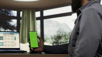 Entrepreneur holding smartphone with greenscreen template while he is examining global revenue trends on laptop. Company CEO connecting on device to help employees, using isolated copyspace. photo