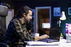 Male programmer works on his portable computer, managing the database and monitoring network for cyber security. Young man sitting at desk and using digital laptop for coding. photo