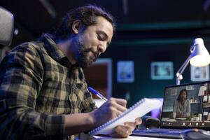 Closeup of bearded man with a pen and notebook, writing meeting notes while on call on the laptop. Male individual positively working from home, using his notepad and portable computer. photo