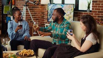 African american man fascinating mates at apartment party with absorbing story. Guest making multiracial group of people laugh in cozy house while drinking alcohol and eating snacks photo