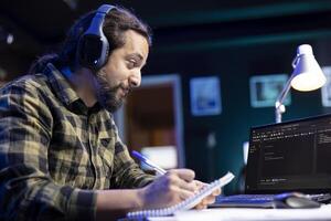 Man with notepad is working on laptop, programming code while listening to music on wireless headset. Male freelancer with headphones using monitoring system software on device and checking his notes. photo