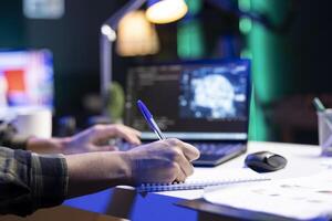 Male individual typing, and computing code for creating databases while writing on his notebook. Closeup of person working from home, monitoring software system and data center. photo