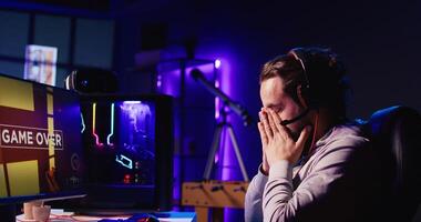 Man upset after losing while playing FPS videogame, using headphones mic to talk with friends. Gamer shutting down computer in anger after receiving game over screen, blaming online teammates photo