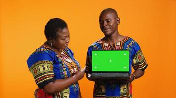 African american partners holding laptop with greenscreen on camera, presenting isolated display with chromakey and standing over orange background. Man and woman showing blank mockup. photo