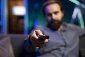 Close up shot of man in neon lit room browsing though movies available on streaming service, enjoying time off from work. Focus on remote control held by viewer in blurry background at home on couch photo