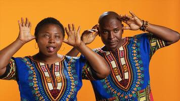 Silly ethnic couple fooling around on camera with comic face, having fun and sticking tongue out in studio. Cheerful people doing funky gesture and enjoying time on set, orange background. photo