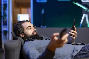 Homeowner bored at home laying down, using TV to watch movies on streaming services and chatting with friends. Apathetic man lounged on couch, mindlessly consuming entertainment and using mobile phone photo