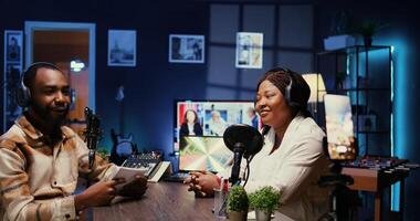 African american host using smartphone on tripod to record entertaining discussion with woman during livestream in apartment studio. Presenter uses mobile phone camera to produce comedy podcast photo