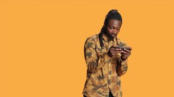 African american guy having fun with videogames online on mobile device, playing rpg contest standing over orange background. Cheerful gamer enjoying gaming tournament in studio. photo