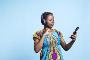Female model playing music from a playing in the studio, having fun with groovy music on audio headset over blue background. Smiling confident woman listening to tunes on headset. photo