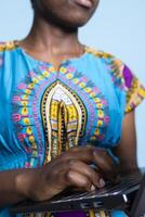 African american girl using mobile pc to scroll across webpages on blue background. Young person with traditional regional attire accesses social networking sites on a laptop in studio. photo
