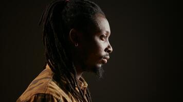 African american guy looking at camera with confidence, feeling happy and relaxed with dreads and cool clothing items. Model posing, standing over black background smiling. Handheld shot. photo