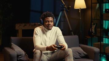 Man in rgb lit apartment playing games on couch, enjoying day off from work. Gamer battling enemies in online multiplayer shooter on gaming console, talking with teammates through headphones photo