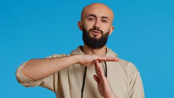 Middle eastern person doing timeout gesture on camera, asking for a break to stop working over blue background. Muslim adult presents symbol to pause or stop something. Camera 2. Handheld shot. photo