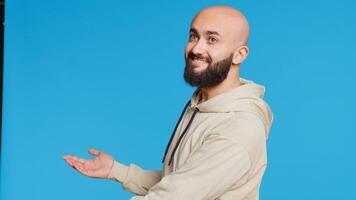 Smiling person advertising something on camera, pointing to left or right sides in studio. Middle eastern guy showing advertisement for marketing campaign, blue background. Camera 2. Handheld shot. photo