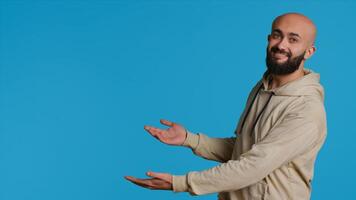 Smiling person advertising something on camera, pointing to left or right sides in studio. Middle eastern guy showing advertisement for marketing campaign, blue background pose. Camera 1. photo