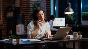 Woman answers phone call from husband while working in office at computer desk. Employee using smartphone to chat with friend during telephone call while at work in front of laptop at night, camera B photo