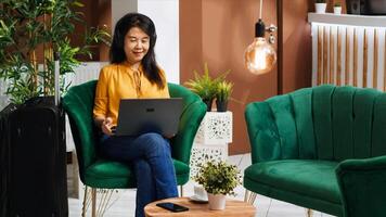 Asian tourist ordering coffee cup while she works on laptop, waiting for check in process in lounge area lobby. Woman arriving early at hotel, relaxing in foyer with refreshment. photo