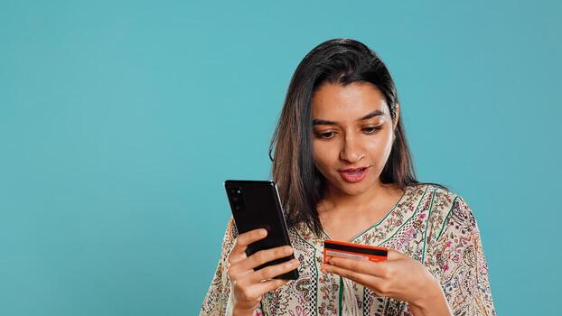 Indian woman excited to purchasing stuff, adding payment method on mobile phone website, isolated over studio background. Ecstatic client does internet shopping, typing debit card info, camera B photo