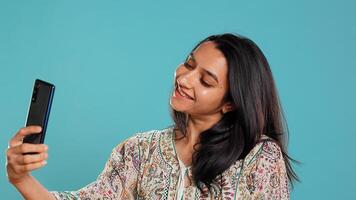 Jolly indian woman smiling for selfies, posting them on social media using cellphone. Joyous girl taking pictures using phone selfie camera, isolated over studio background, camera B photo