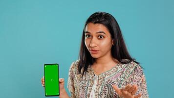 Woman presenting green screen mobile phone, isolated over studio background. Cheerful indian person holding copy space chroma key smartphone used for advertising brands, camera B photo