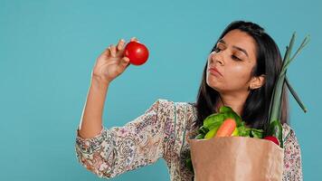 Woman with recycled paper bag in hands filled with vegetables testing quality, studio background. Sustainable living person inspecting groceries after doing purchases in shop, camera B photo