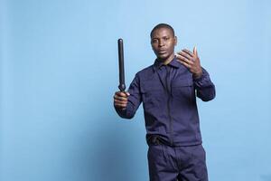 Security guard with a baton in his hand calling people over, asking persons to approach him against blue background. Young armed patrol agent working on public protection and safety. photo