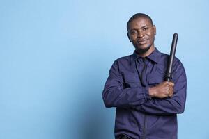 African american security officer feels proud with his duty in studio, posing with confidence holding his baton dressed in professional uniform. Skilled bodyguard standing against blue background. photo