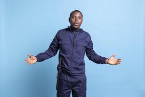 Security guard in uniform is acting surprised in front of the camera, being in shock after finding out something. African american protection agent with baton feels amazed against blue background. photo