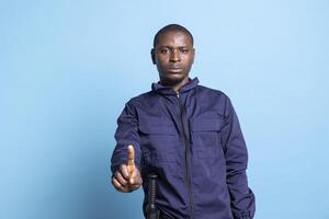 African american security guard poses with a finger up in studio, presenting a restriction sign against blue background. Person working as a patrol officer showing a stop symbol in studio. photo