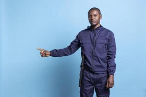 African american bodyguard pointing to something aside in studio, working on an advertisement against blue background. Male security officer in uniform indicates a left or right direction. photo