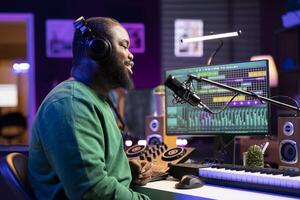 Male sound engineer singing a new song to record audio on microphone, editing music with mixing console and gear in home studio. Artist adjusting volume levels with stereo tools. photo