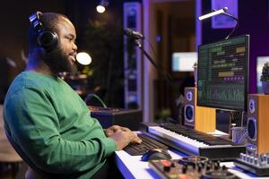 African american composer mixing and adding sounds on his recordings, listening to the final product on headset. Audio engineer producing new music, editing volume levels in home studio. photo