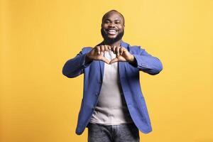 Portrait of loving joyous african american man doing heart symbol shape with hands. Happy caring BIPOC person showing love gesturing, isolated over yellow studio background photo