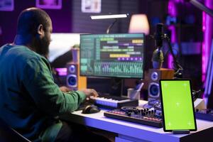 Composer editing his audio files and recordings on daw software, sitting next to a greenscreen display. Skilled sound technician uses mixing and mastering gear to produce new material. photo