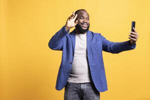 Cheerful man using smartphone to take selfies and post them on social media. Happy BIPOC person taking photos using phone selfie camera, isolated over yellow studio background