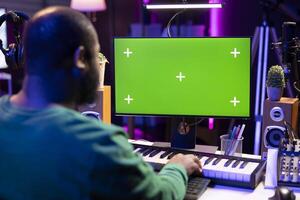 Producer composing music with electronic keyboard notes and greenscreen display on pc, post production in home studio. Musician artist plays midi controller next to mockup layout. photo