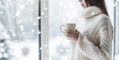 Caucasian woman in a cozy white sweater holding a steaming mug by a window. Concept of warmth winter serenity and relaxation. Banner. Copy space. Christmas, New Year photo