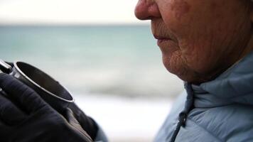 A woman is standing on a beach holding a mug. She is wearing a white hat and a blue jacket. The scene is calm and peaceful, with the ocean in the background. video