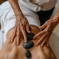 Hot stone massage therapy session, featuring a therapist gently placing stones on a client's back, in a room with soft natural light and white linens. Spa treatment concept photo