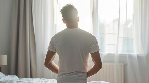 Caucasian man standing by a sunlit window in a bedroom. Concept of solitude, morning routine, introspection, personal space photo