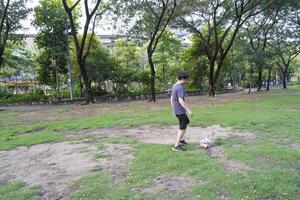 asiático hombre son corriendo y pateando pelota para jugando un fútbol a al aire libre parque. foto