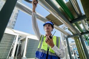 Industrial engineer adjusting pipeline valve with radio communication on construction site. Engineer on urban construction site using radio to coordinate with team on a sunny industrial rooftop. photo