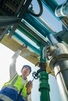 Industrial engineer adjusting pipeline valve with radio communication on construction site. Construction worker using walkie-talkie to coordinate machinery adjustments on a sunny day. photo