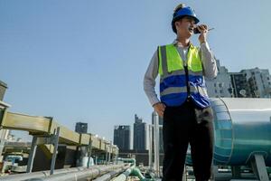 A young engineer checks equipment on a rooftop amidst urban skyline, clipboard in hand, ensuring system functionality.Engineer in safety vest and helmet conducting maintenance on rooftop machinery. photo