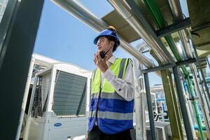 Industrial engineer adjusting pipeline valve with radio communication on construction site. Engineer on urban construction site using radio to coordinate with team on a sunny industrial rooftop. photo