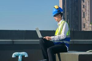 enfocado ingeniero en difícil sombrero y la seguridad chaleco utilizando walkie-talkie y ordenador portátil en construcción sitio con urbano fondo. foto