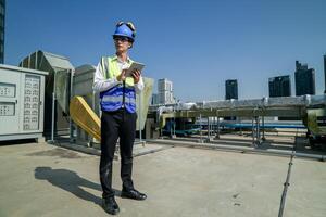 Engineer with tablet on rooftop assessing HVAC system against city skyline, safety equipment and hard hat on. photo